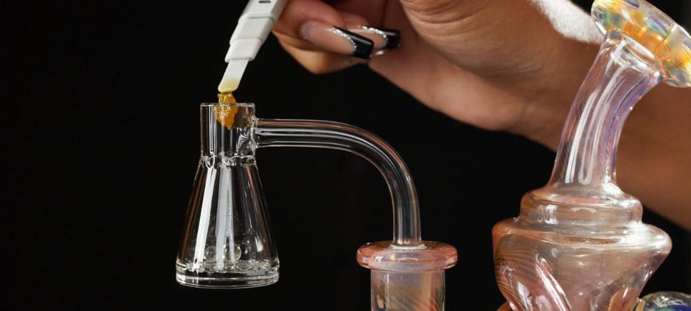 Primer plano de una mano colocando concentrado sobre un banger de vidrio transparente montado en un dab rig, con fondo oscuro.